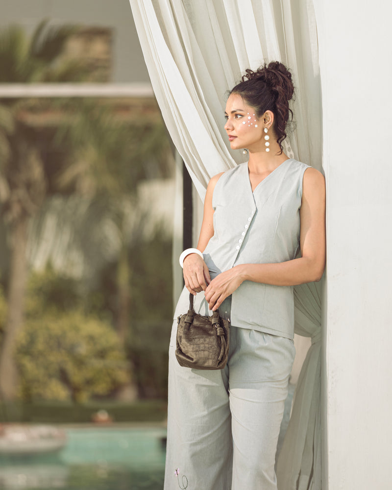 Woman in light gray outfit holding a brown handbag by a window with white curtains.