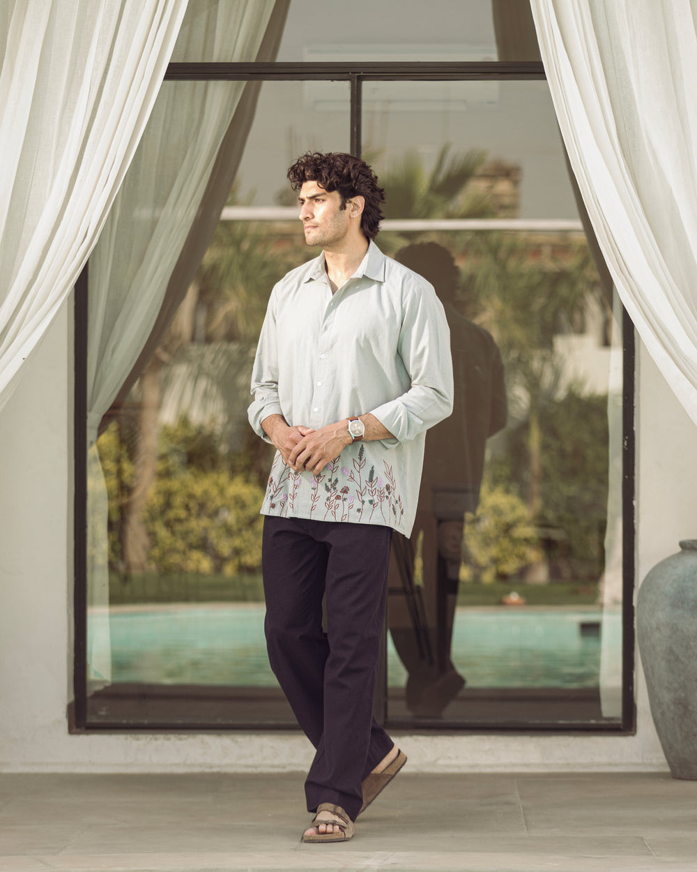 Man standing in a modern indoor setting with large windows and white curtains.

