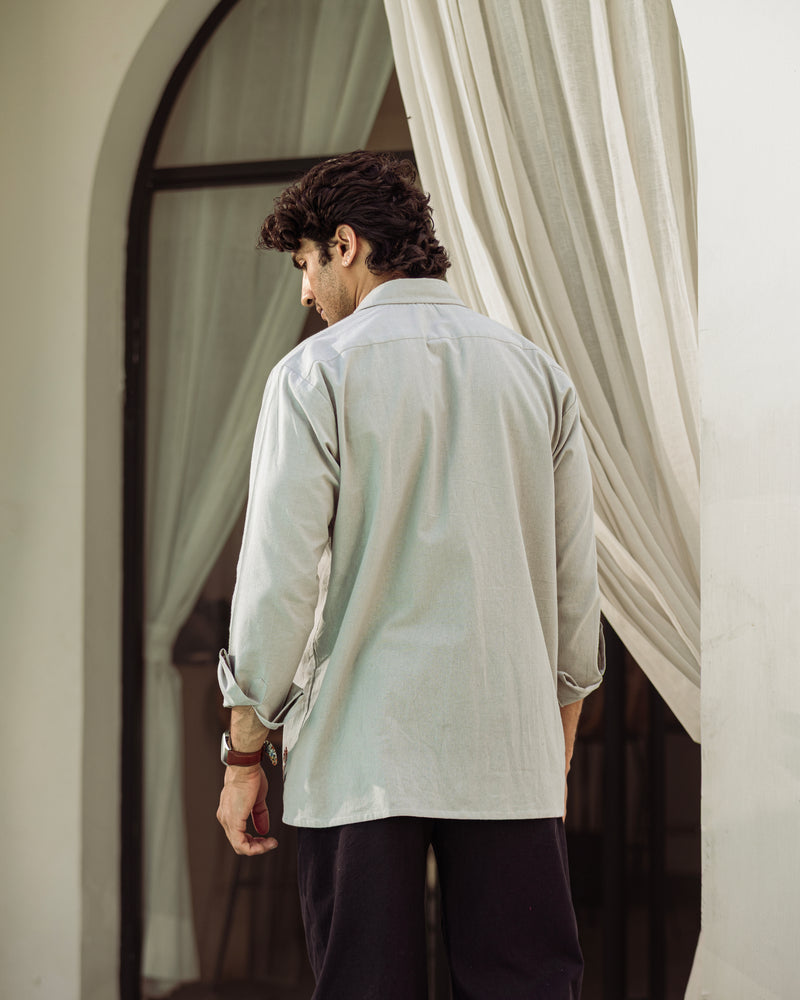 Man wearing a light-colored shirt standing in front of a mirror with white curtains.

