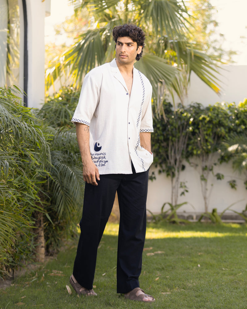 Man wearing a white shirt with black text and dark pants standing outdoors with greenery in the background