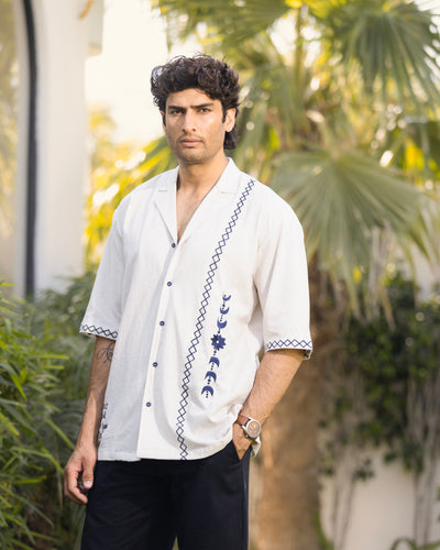 Man wearing a white embroidered shirt with black pants standing outdoors with greenery in the background