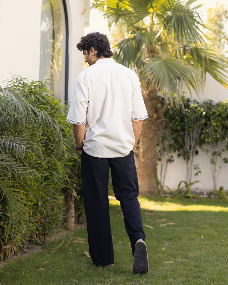 Man in a white shirt and dark pants walking in a garden with greenery.