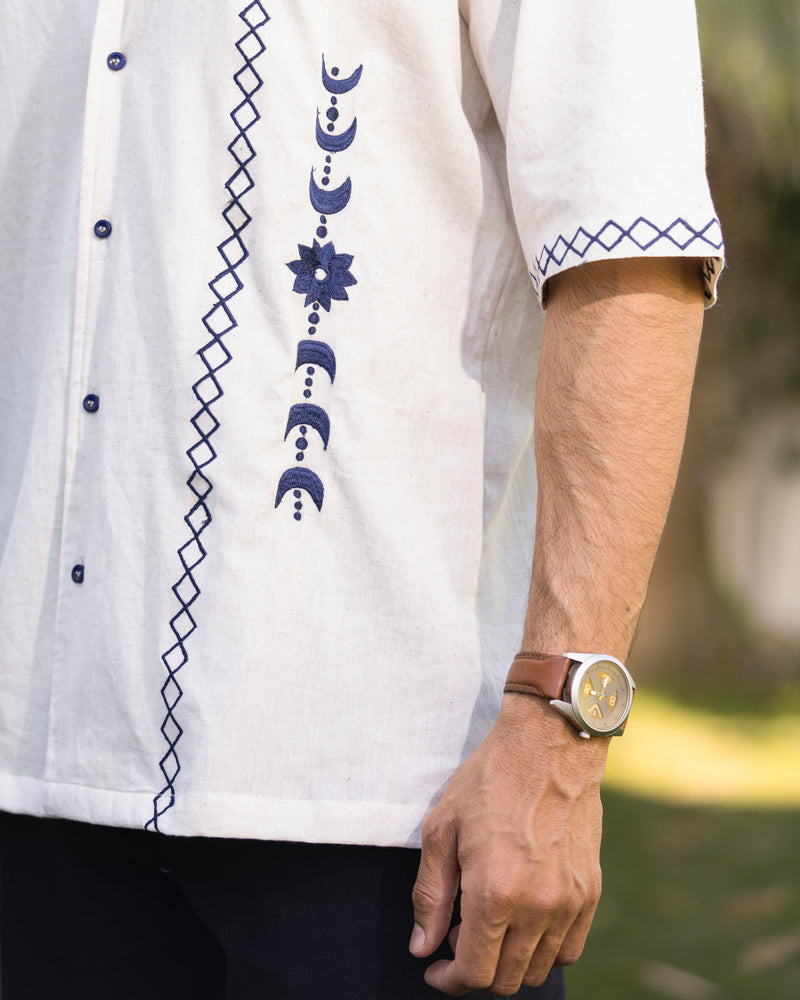 White embroidered shirt with blue patterns worn by a person, with a blurred natural background.