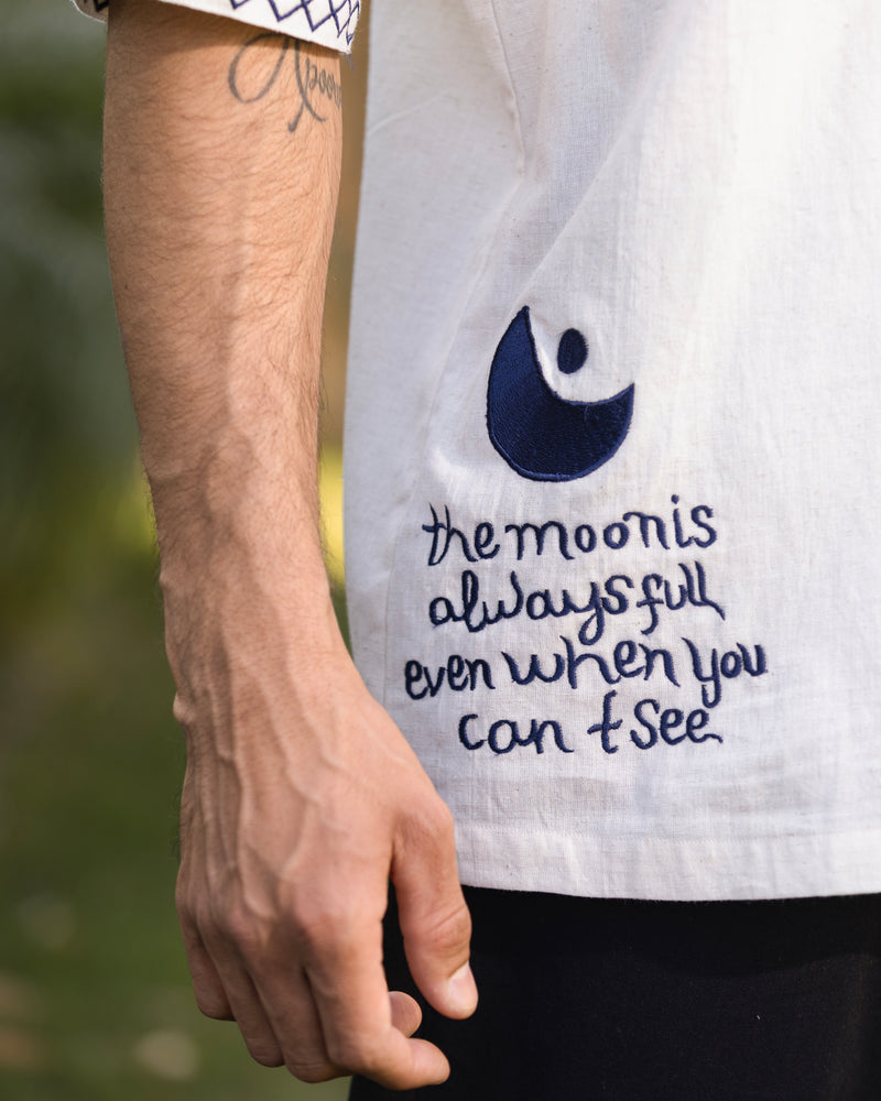 Person wearing a shirt with a moon design and text, holding a blurred natural background.