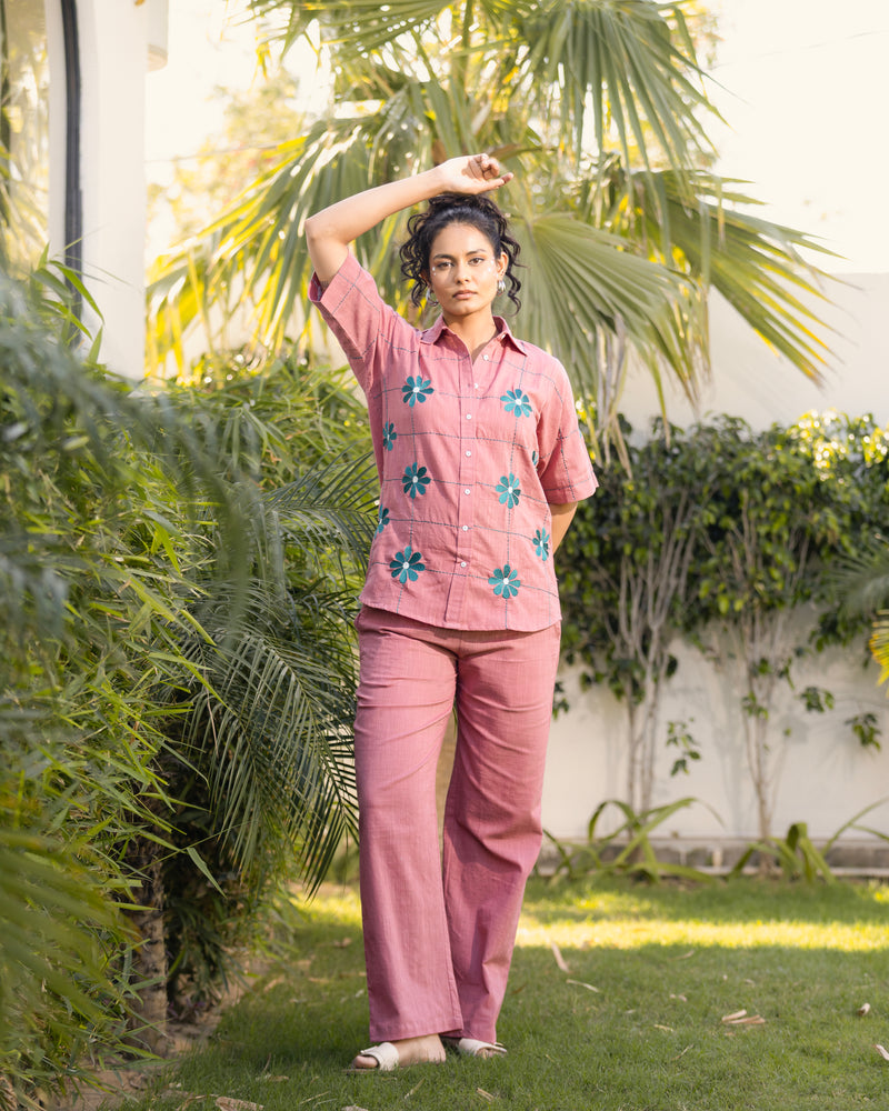 Woman in a pink outfit with floral patterns standing outdoors among greenery.