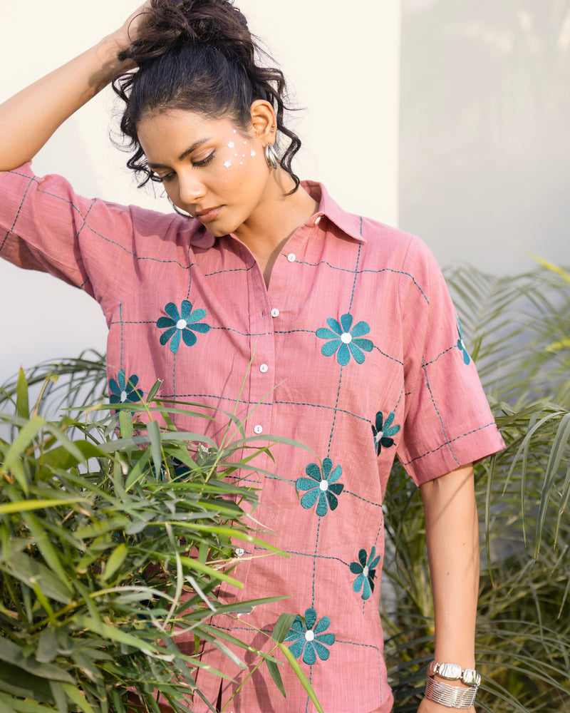 Woman wearing a pink shirt with blue floral patterns, standing among green plants.