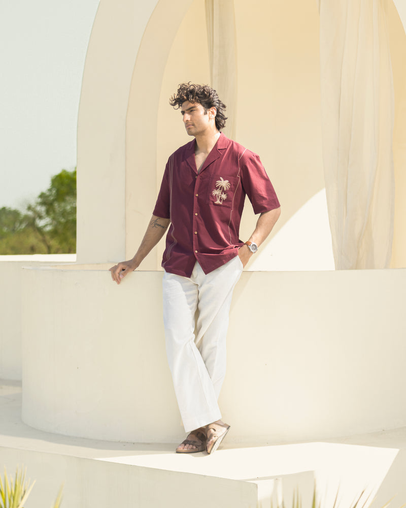 Man in a red shirt and white pants leaning against a white architectural structure with greenery in the background.