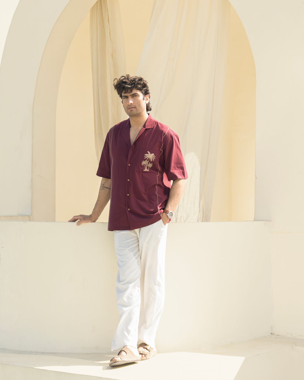 Man wearing a maroon shirt with a palm tree design and white pants standing against a light architectural background.