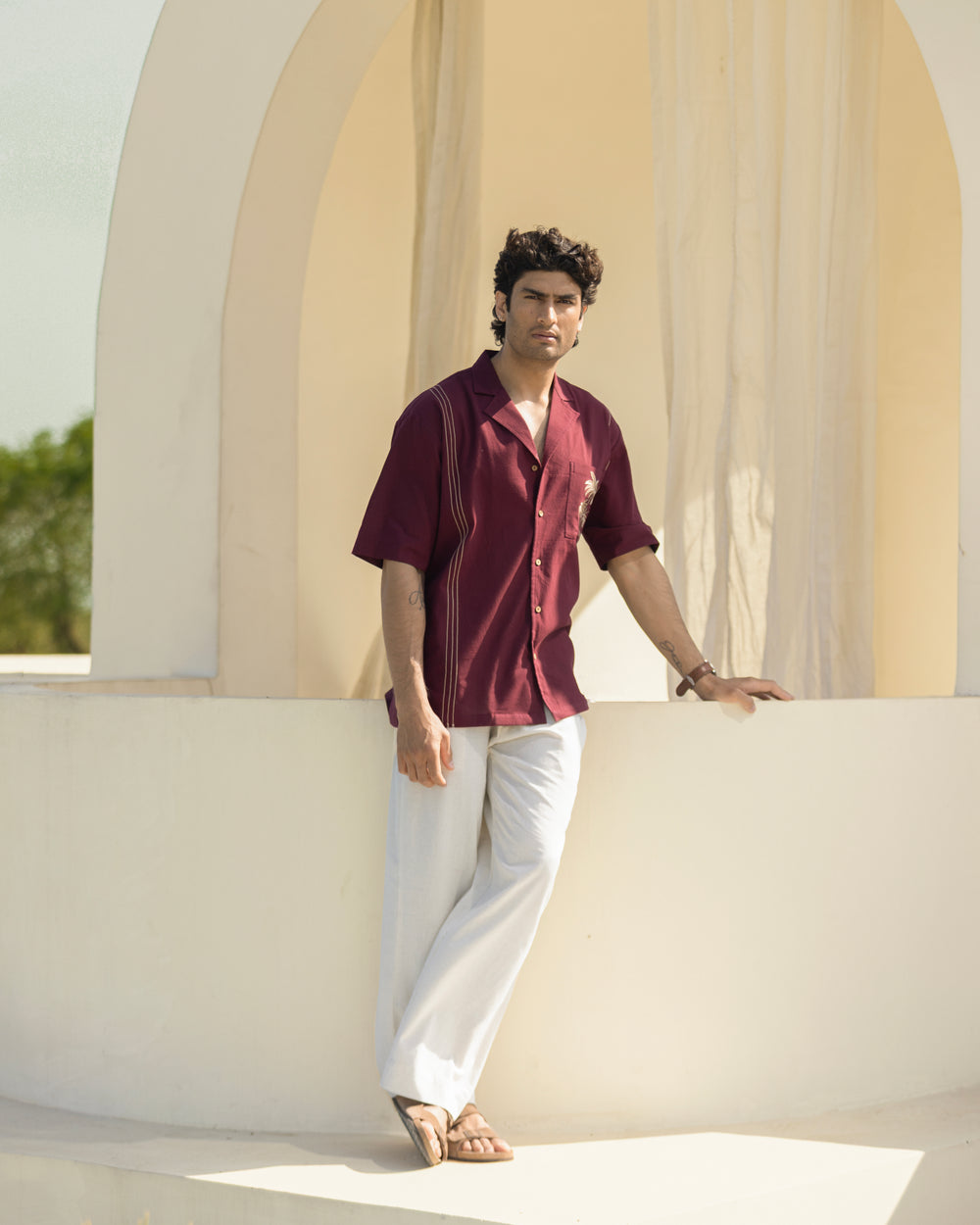Man in a maroon shirt and white pants leaning against a light-colored architectural structure.