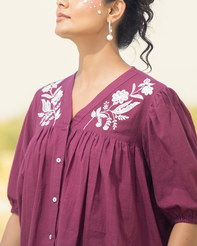 Woman wearing a purple blouse with white floral embroidery against a blurred natural background