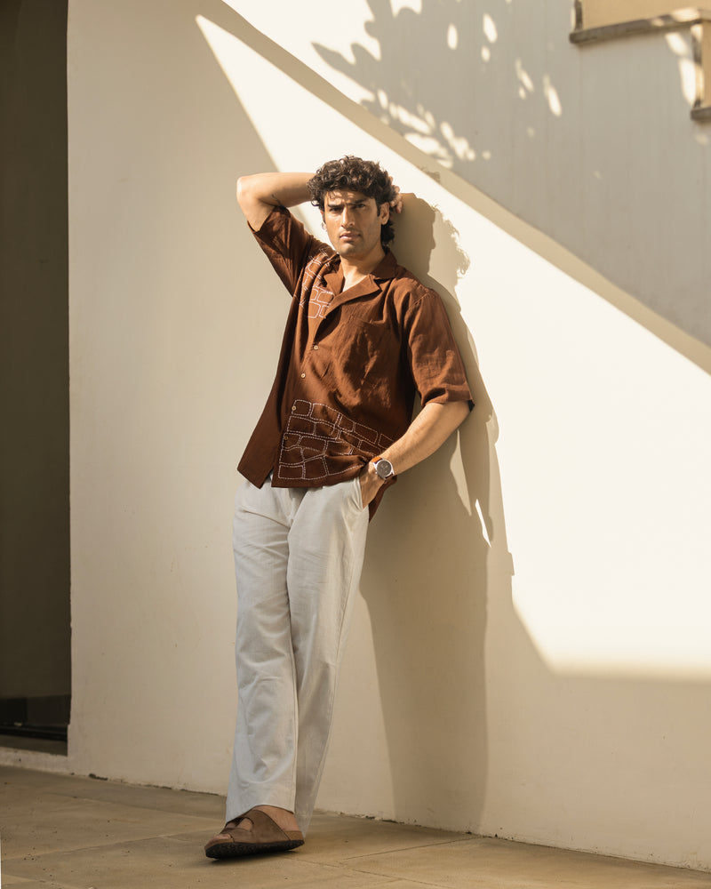 Man wearing a brown shirt and white pants leaning against a wall with sunlight casting shadows.