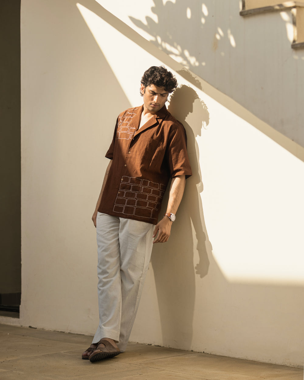 Man wearing a brown patterned shirt and white pants leaning against a light-colored wall.