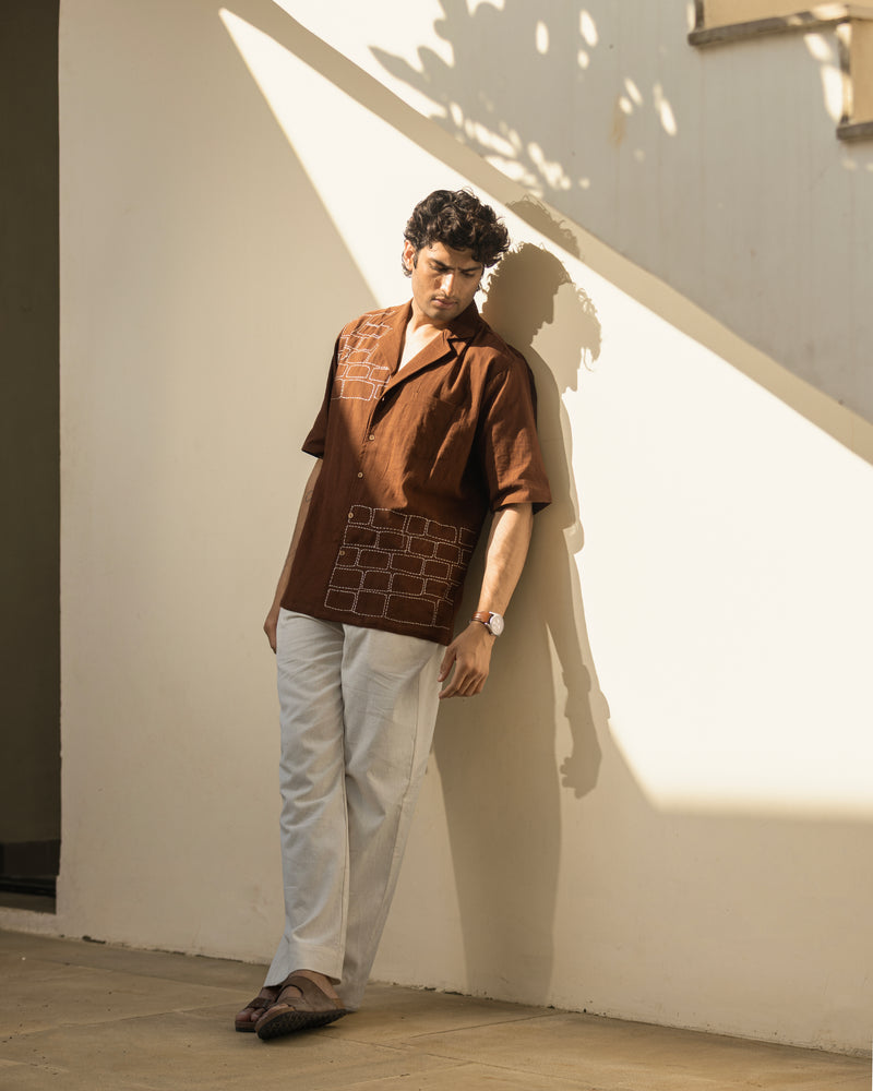 Man wearing a brown patterned shirt and white pants leaning against a light-colored wall.