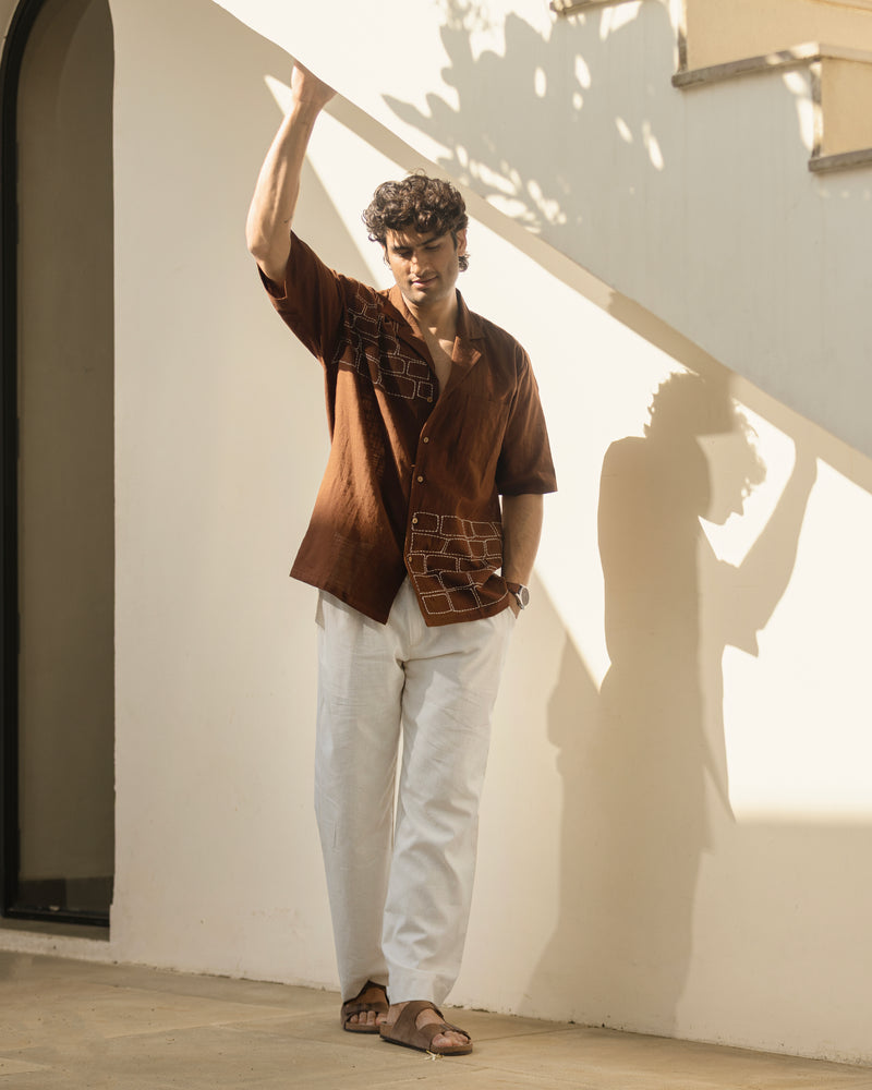 Man wearing a brown shirt and white pants standing against a light-colored wall.