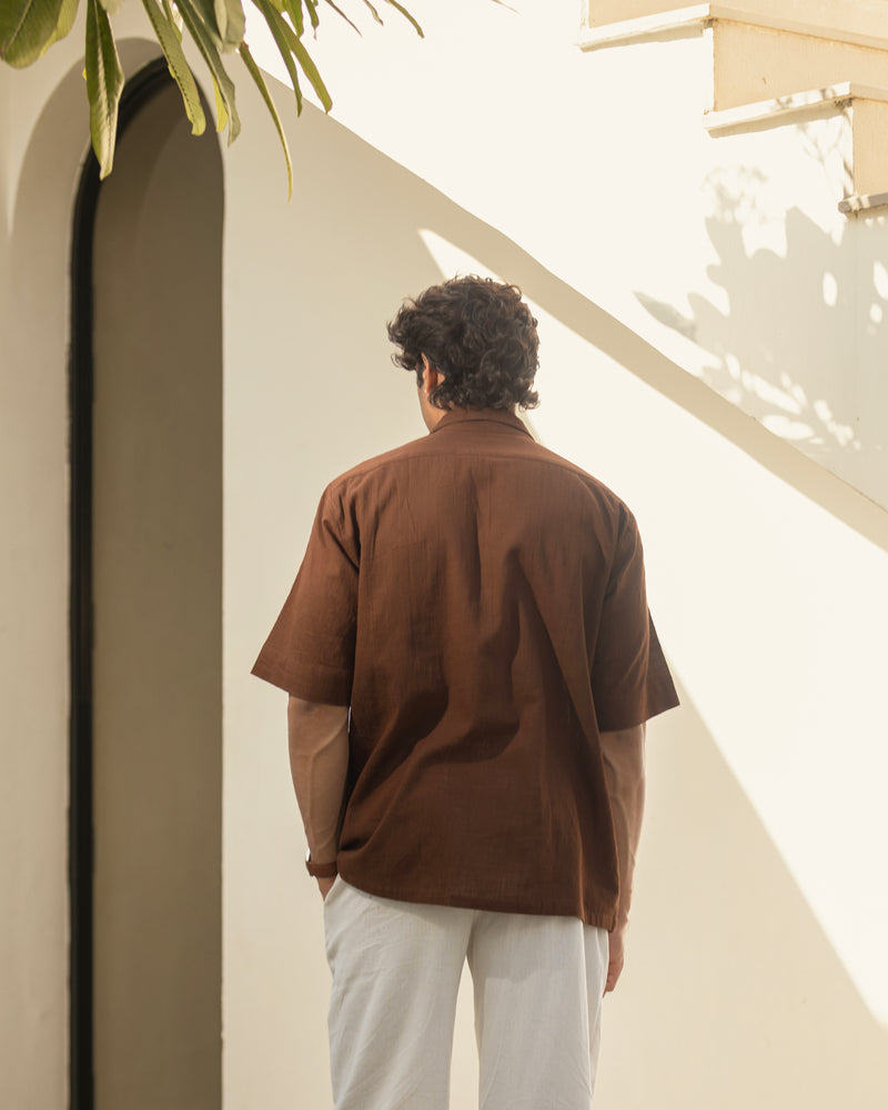 Person wearing a brown shirt and light-colored pants standing in a sunlit outdoor area.