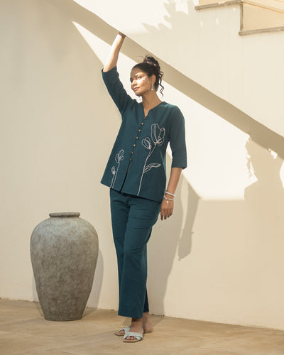 Woman in a teal outfit standing next to a large vase in a room with wooden flooring and white walls.