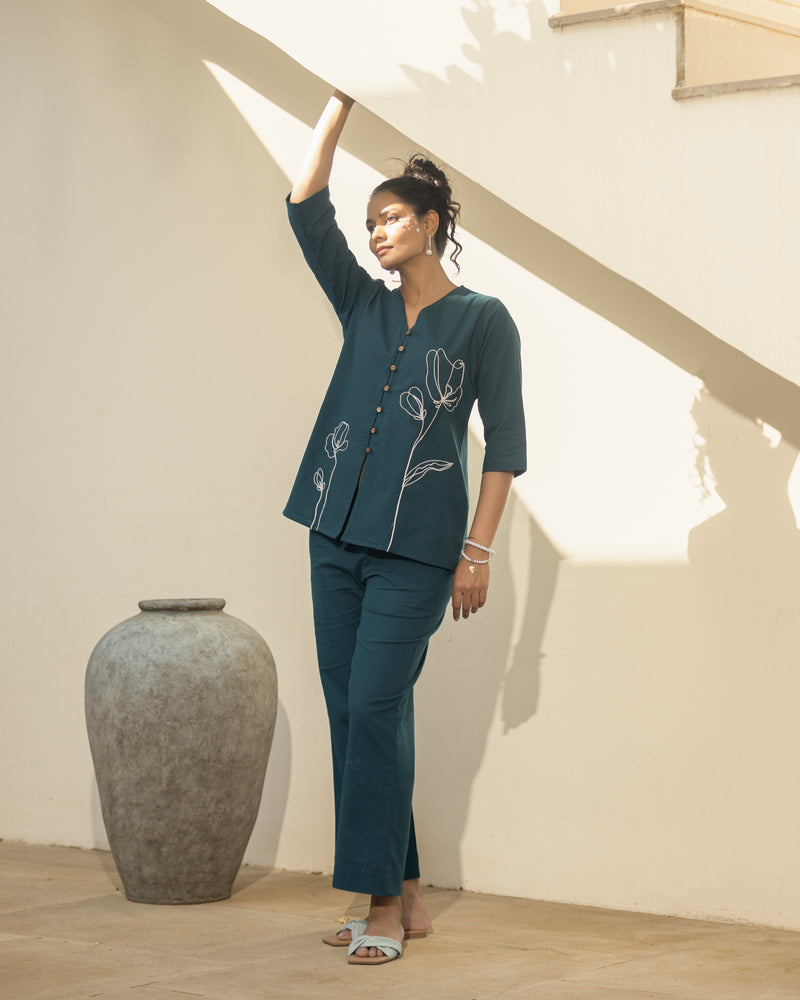 Woman in a teal outfit standing next to a large vase in a room with wooden flooring and white walls.