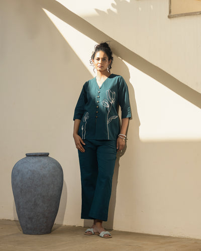 Woman in teal outfit standing next to a large vase in a sunlit room
