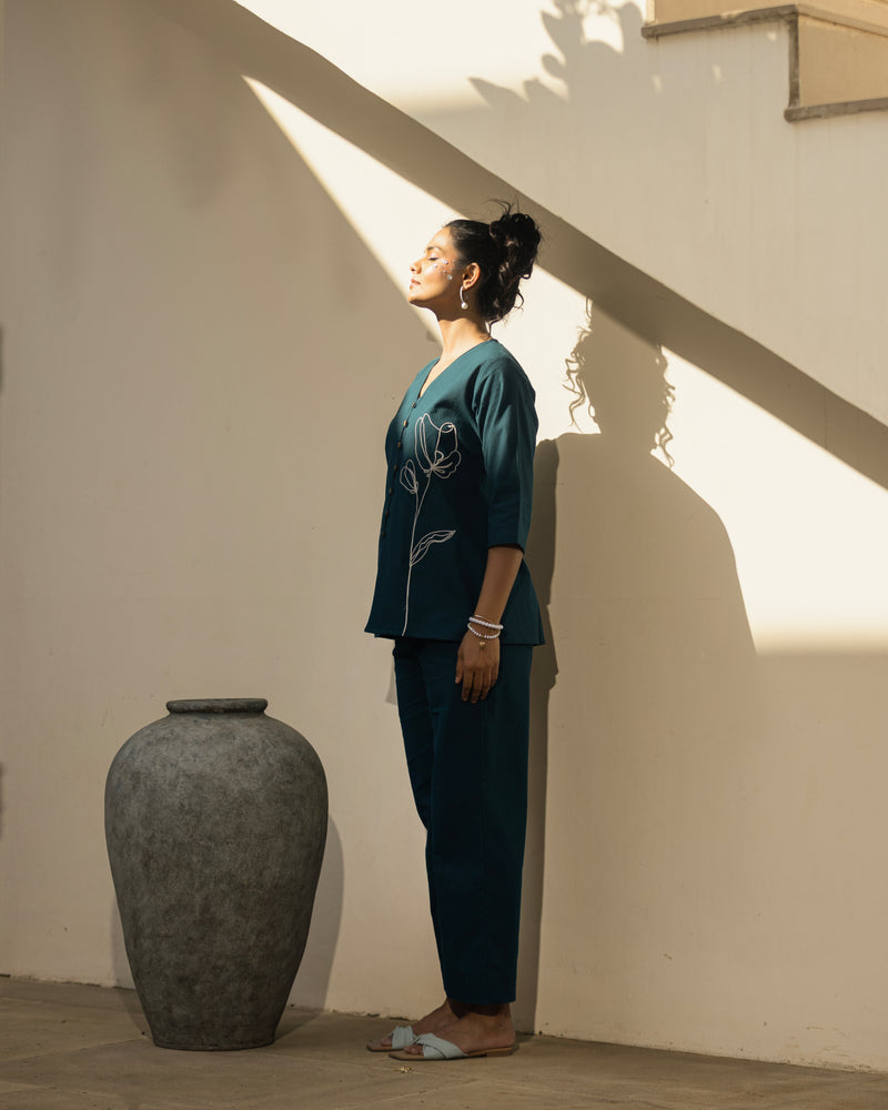 Woman in a teal outfit standing next to a large vase in a sunlit room.