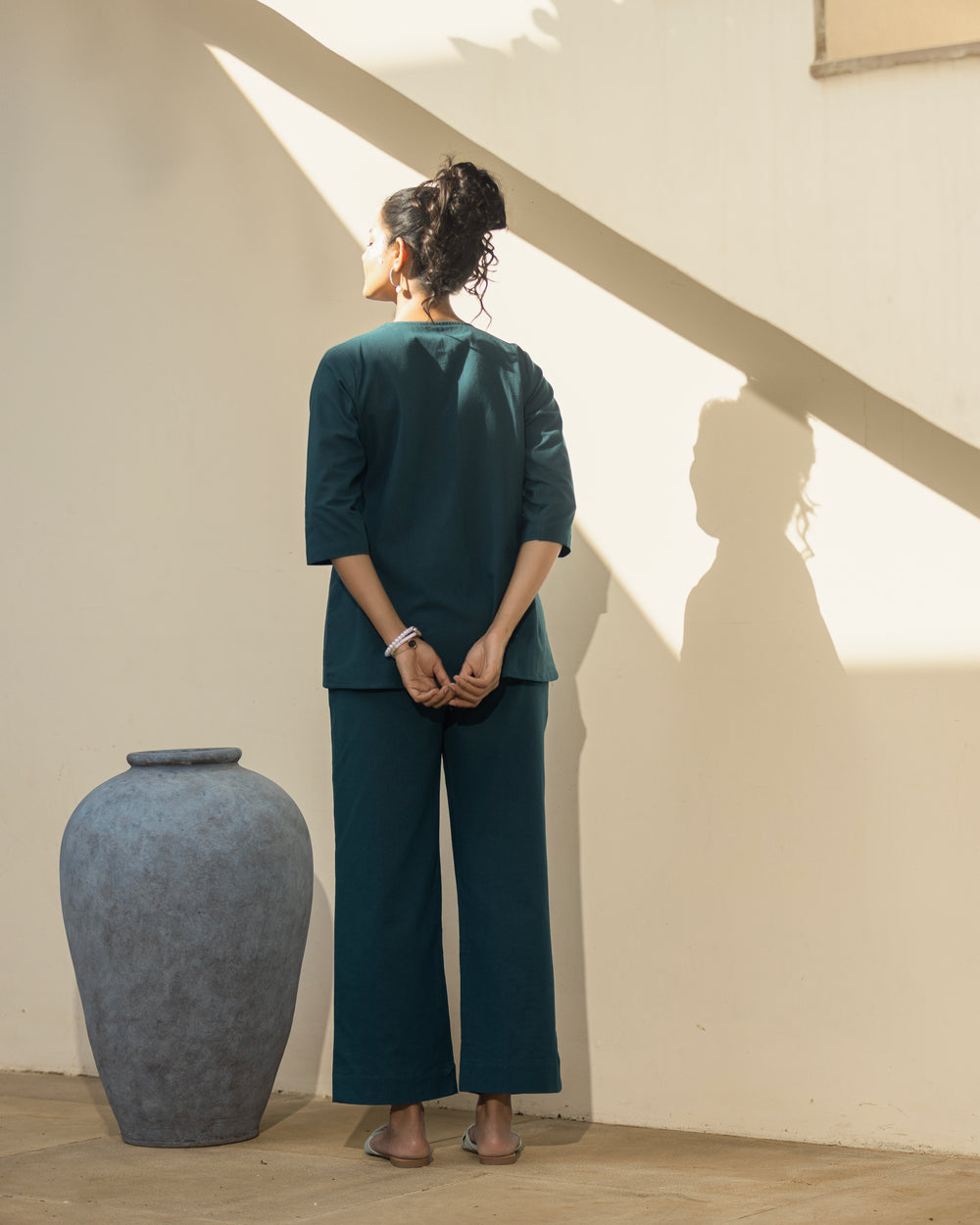 Person wearing a teal outfit standing in a room with a large vase and sunlight casting shadows on the wall.