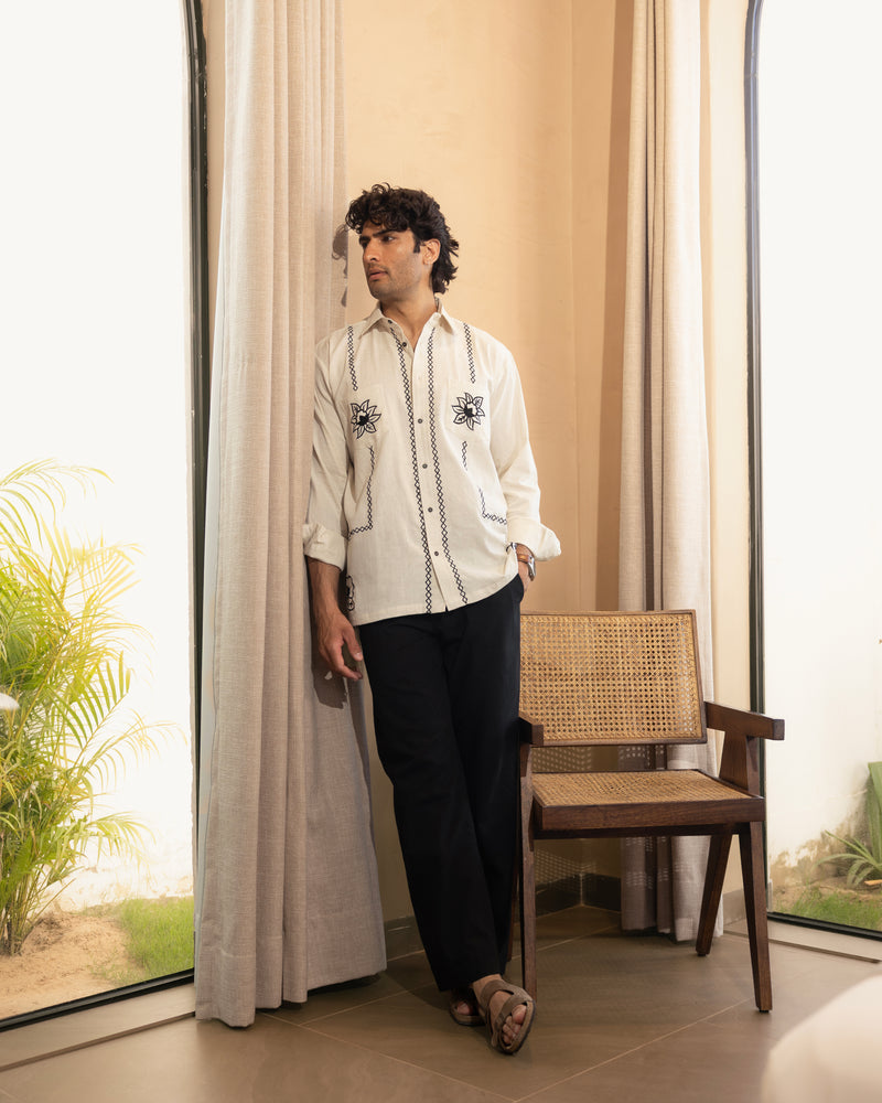 Man standing in a room with large windows and curtains, wearing a white embroidered shirt and dark pants.