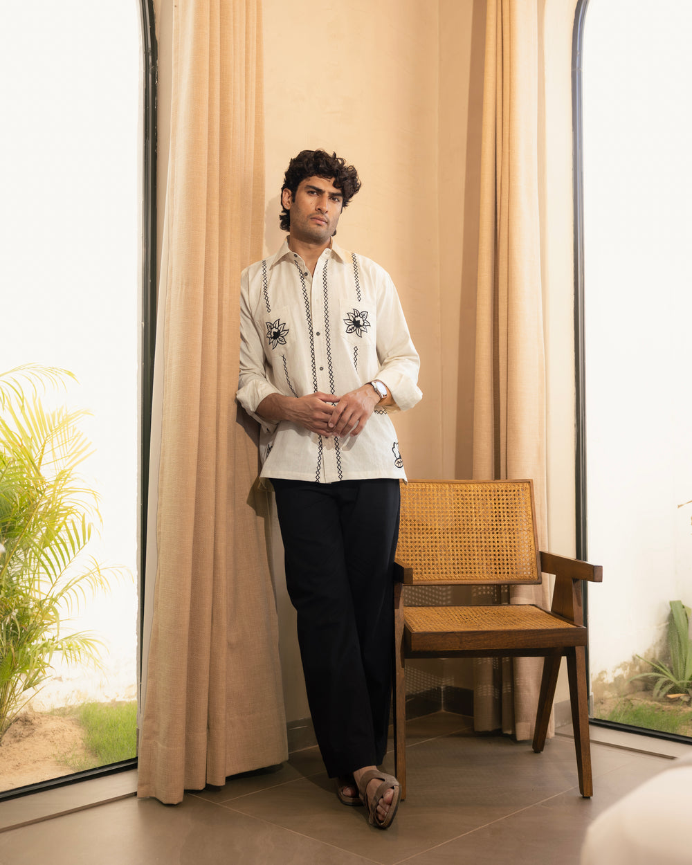 Man in a white embroidered shirt and black pants standing in a room with beige curtains and a wooden chair.