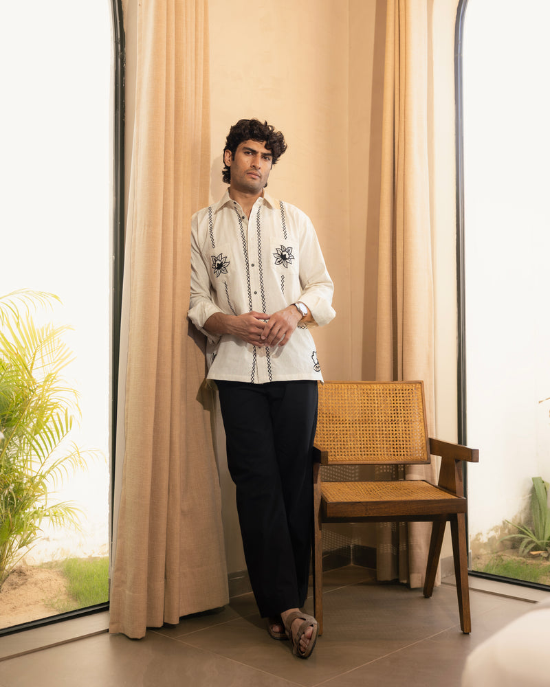Man in a white embroidered shirt and black pants standing in a room with beige curtains and a wooden chair.