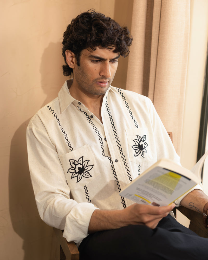 Man reading a book wearing a white embroidered shirt indoors.