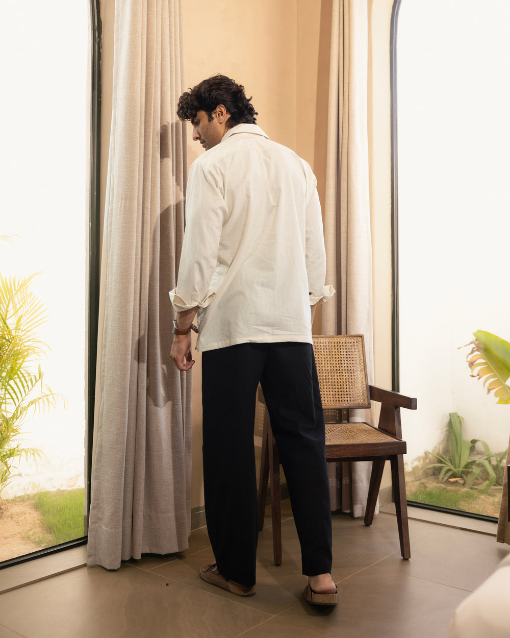 Man standing in a room with large windows and curtains, wearing a white shirt and black pants.