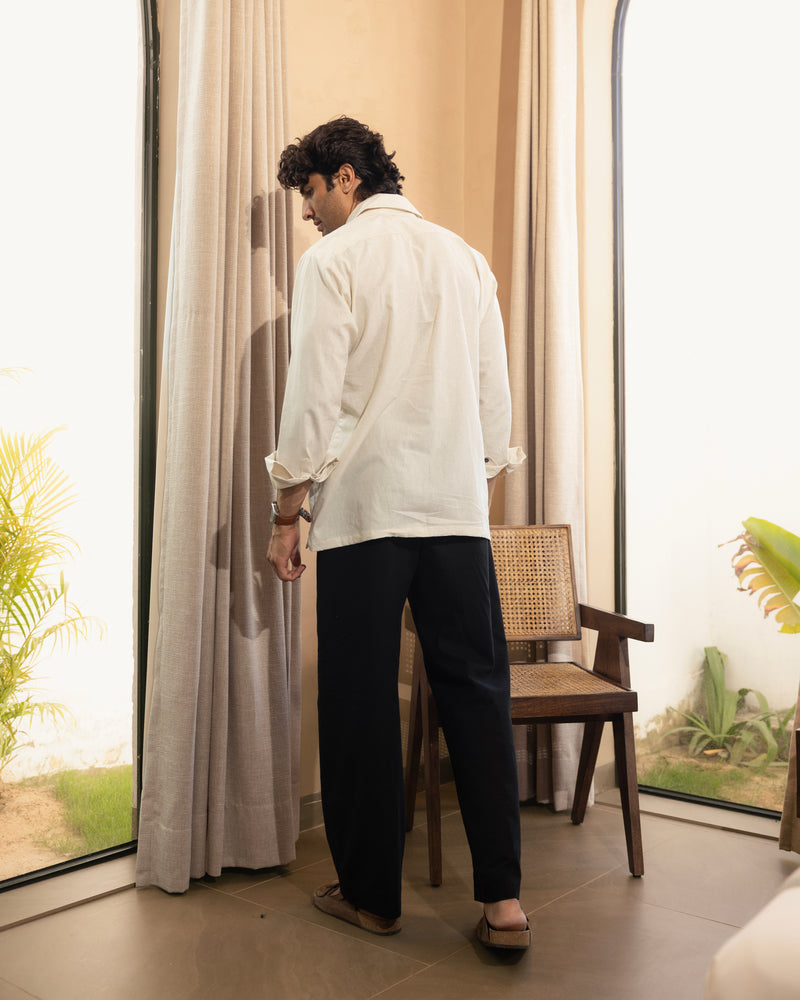 Man standing in a room with large windows and curtains, wearing a white shirt and black pants.