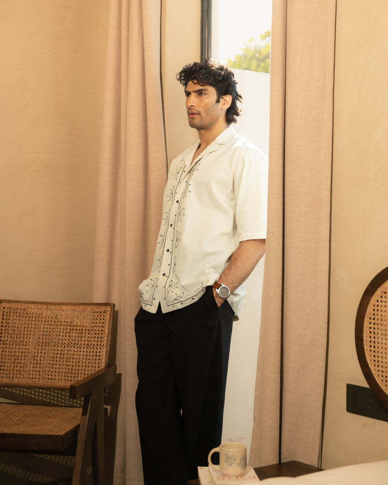 Man standing in a room with beige curtains and a wicker chair.

