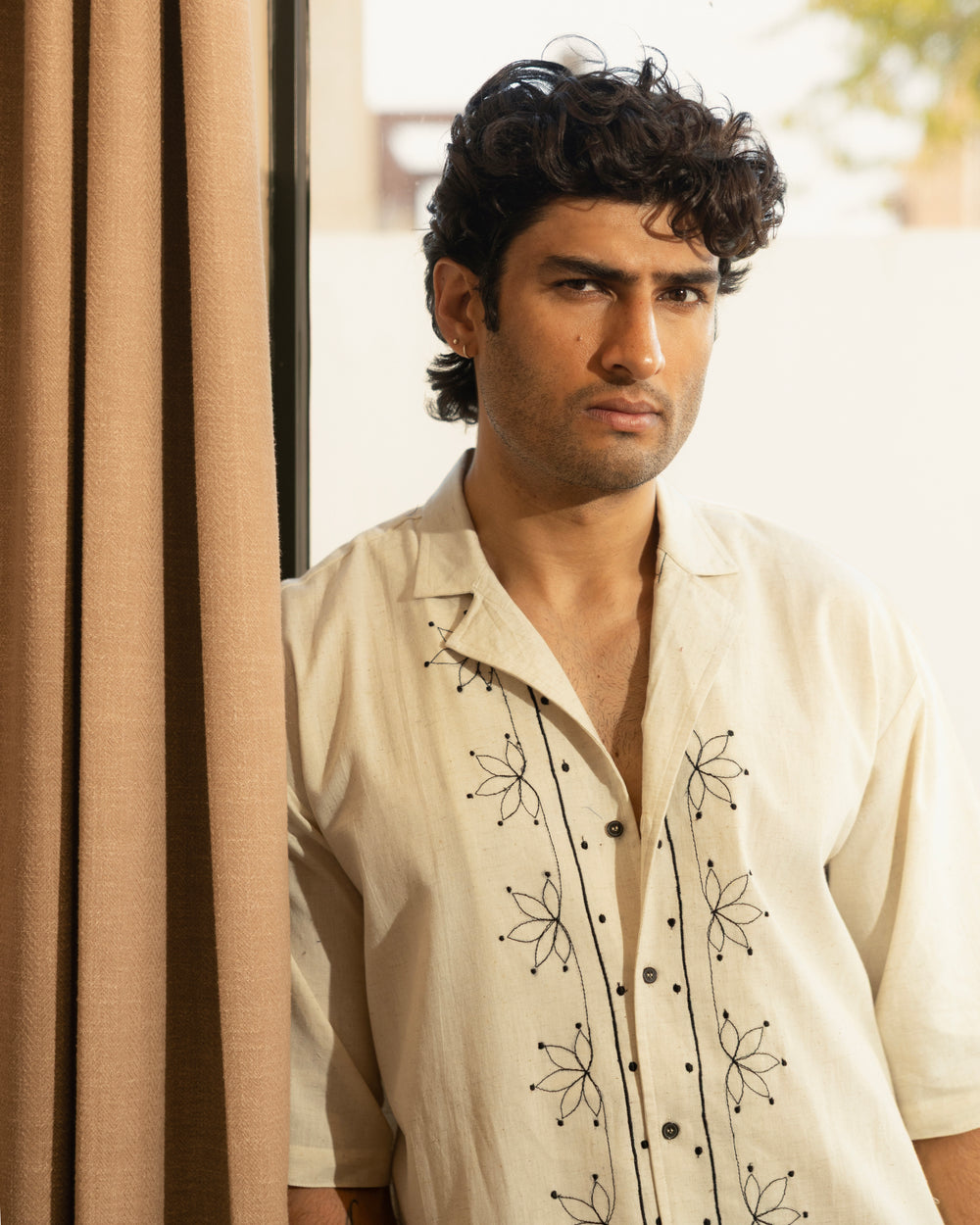 Man wearing a cream-colored shirt with black floral embroidery standing against a beige curtain.

