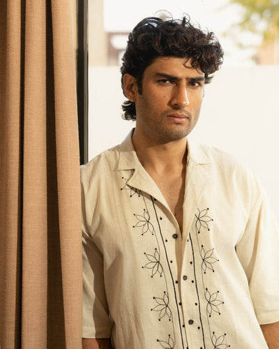 Man wearing a cream-colored shirt with black floral embroidery standing against a beige curtain.

