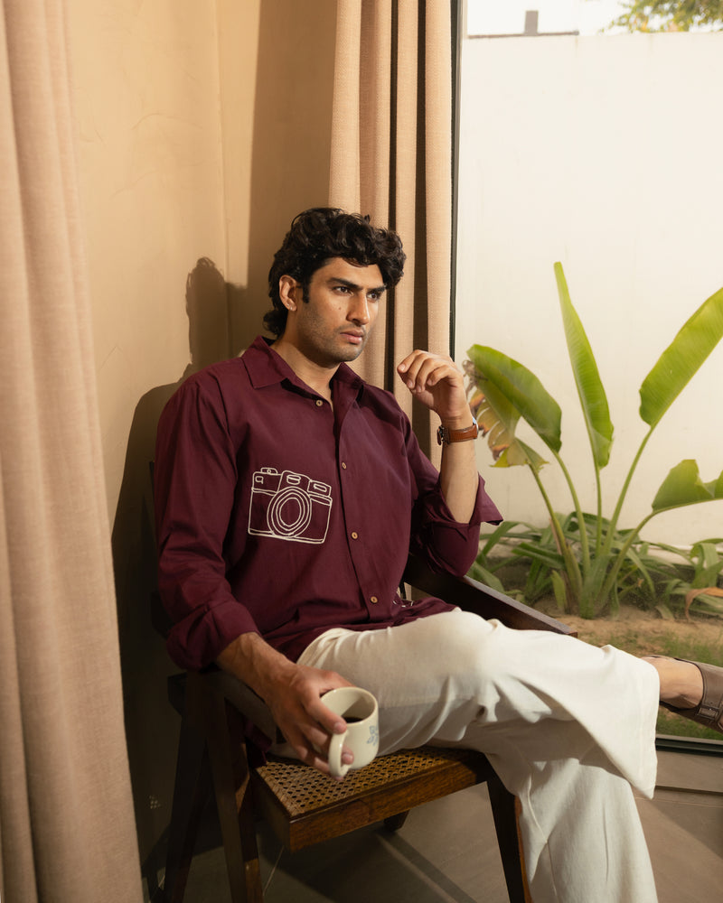 Man sitting in a chair holding a cup, wearing a maroon shirt with a camera design and white pants.

