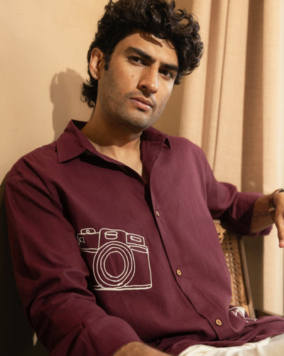 Man wearing a maroon shirt with a camera design, sitting indoors.

