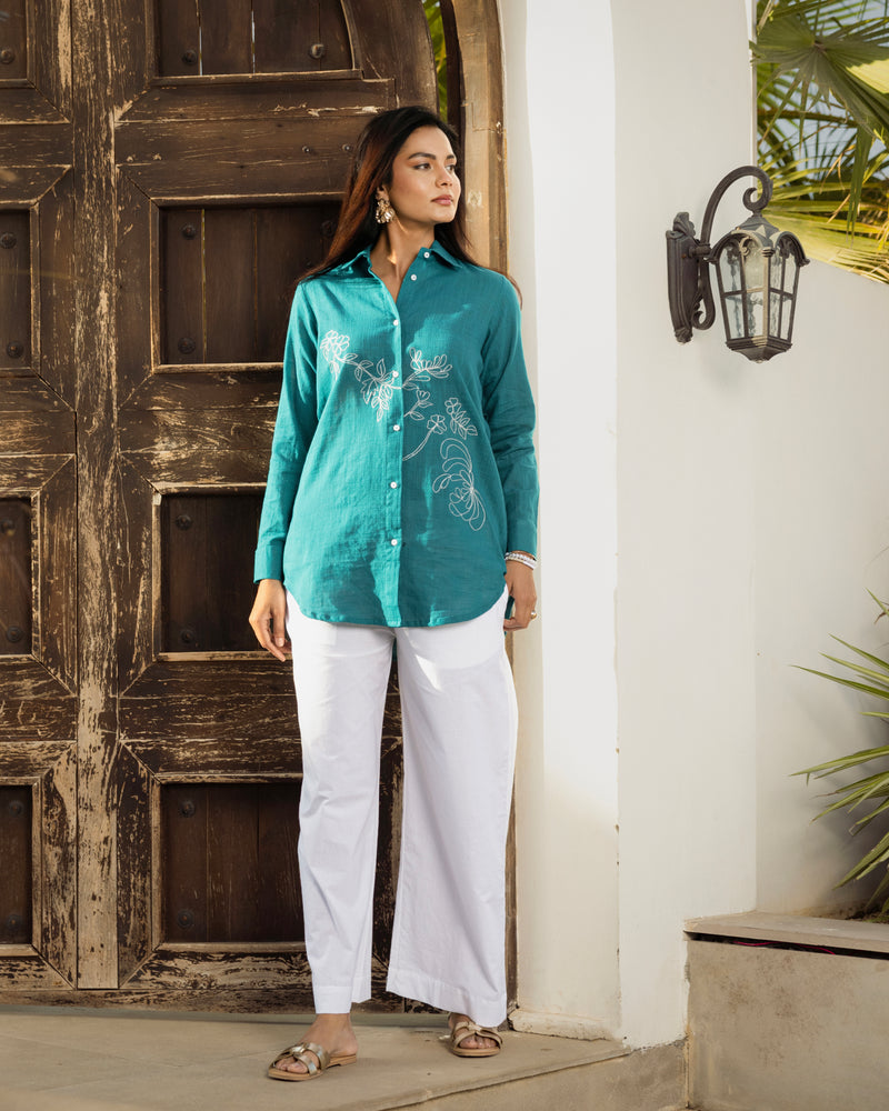 Woman wearing a teal shirt with white pants standing in front of a wooden door.