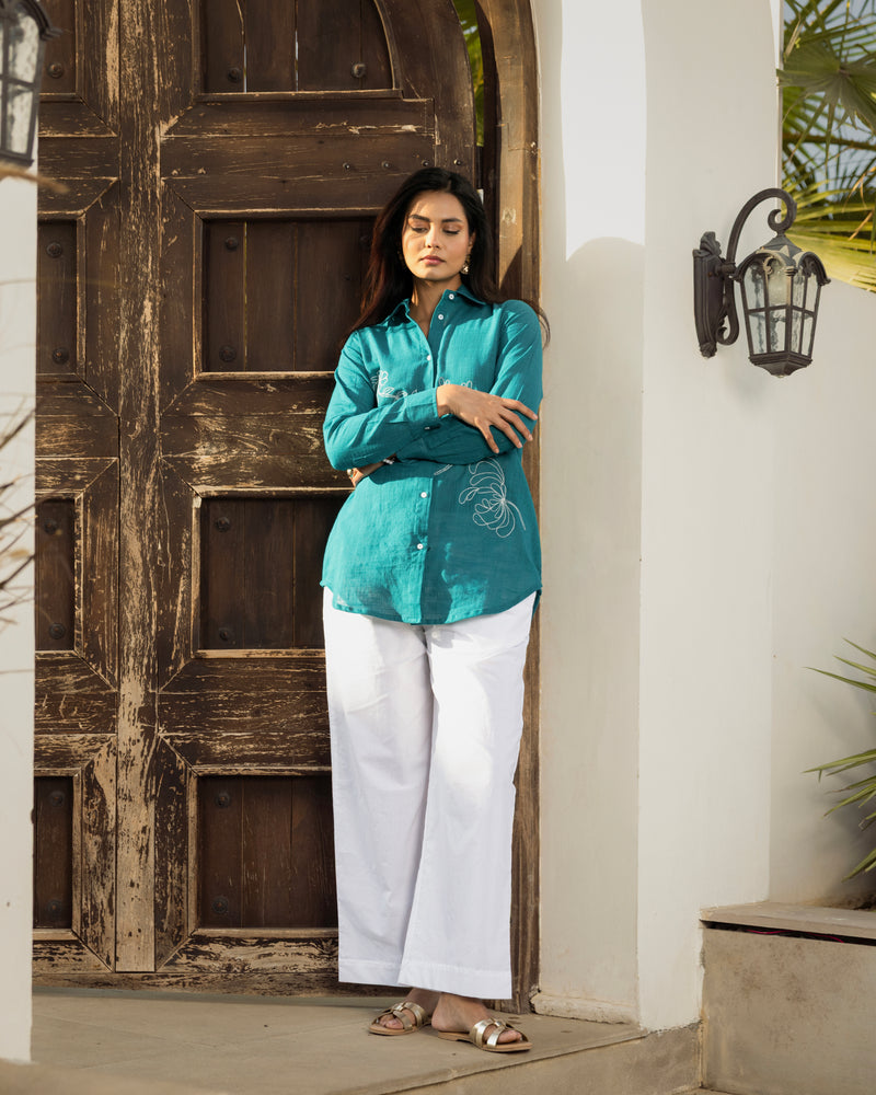 Woman in a teal shirt and white pants standing in front of a wooden door.