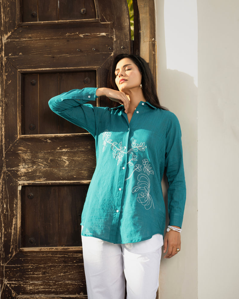 Woman wearing a teal embroidered shirt and white pants standing in front of a wooden door.