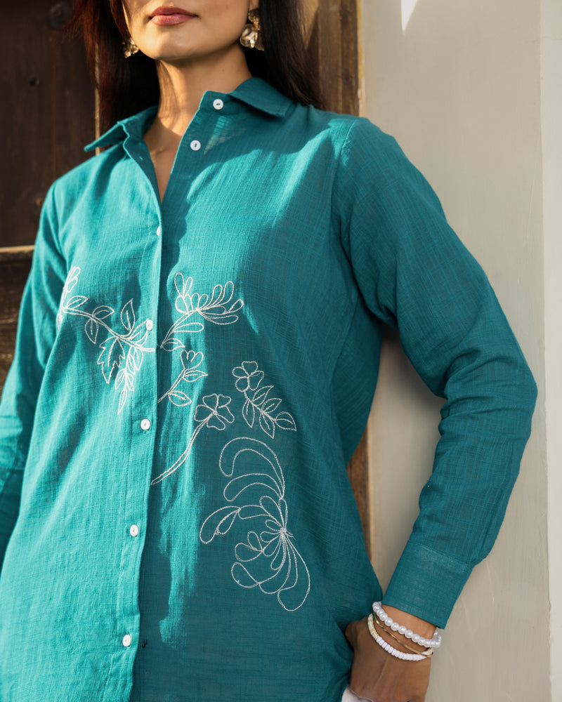 Teal shirt with white floral embroidery worn by a person against a neutral background