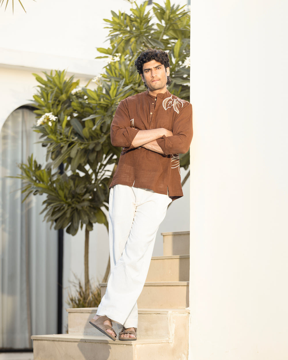 Man in a brown shirt and white pants standing on steps with greenery in the background