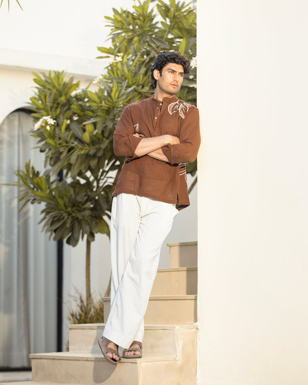 Man in a brown shirt and white pants standing on steps with greenery in the background