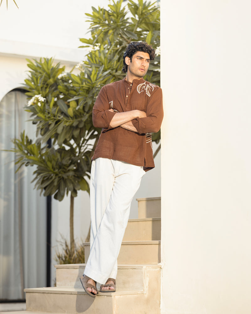 Man in a brown shirt and white pants standing on steps with greenery in the background