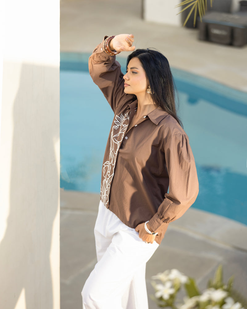 Woman in a brown blouse with white embroidery and white pants standing by a pool.