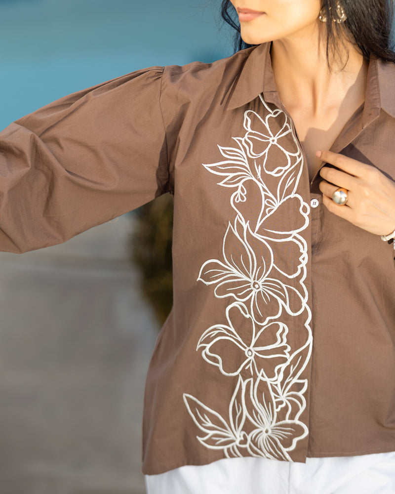 Brown shirt with white floral embroidery worn by a person against a blurred background