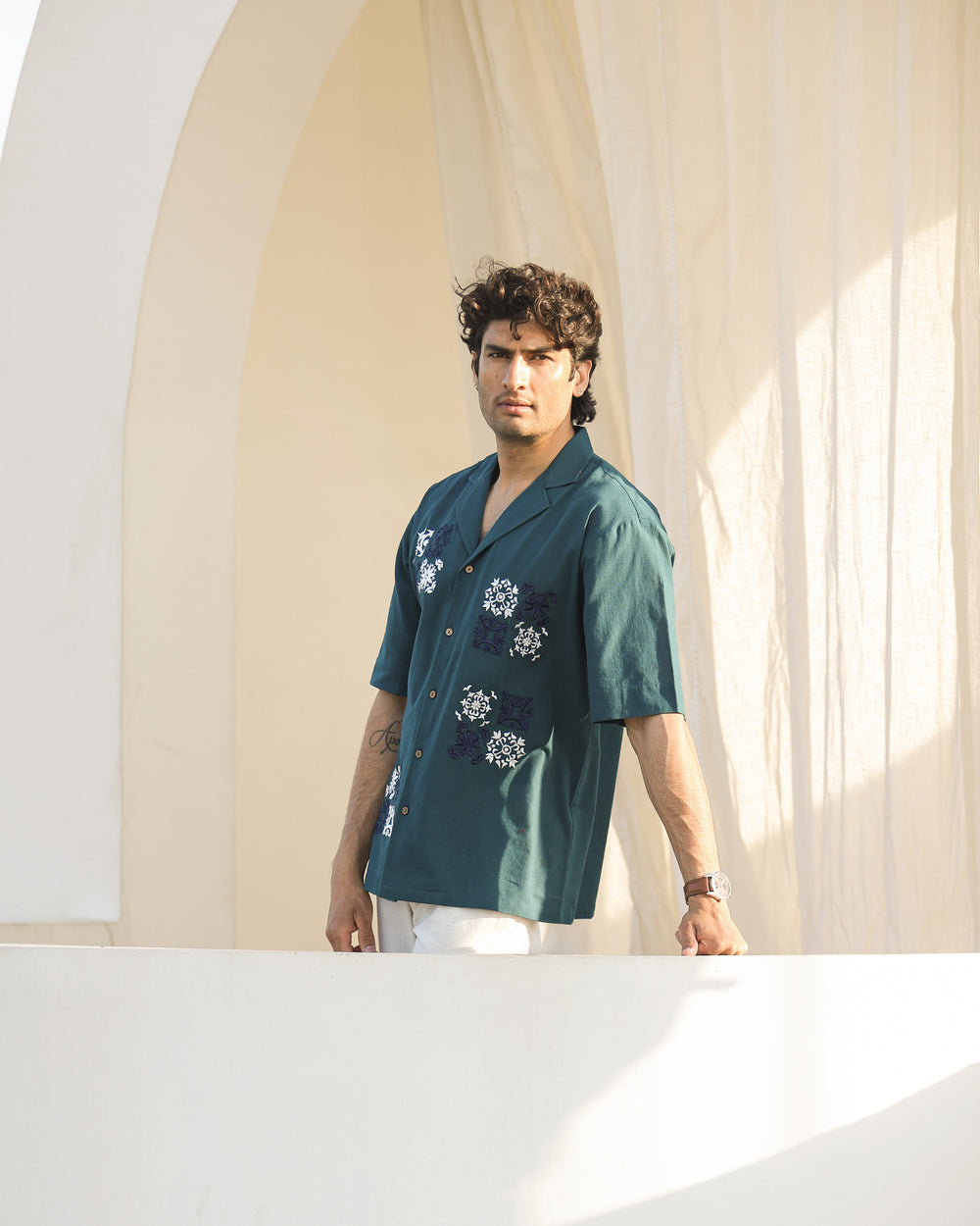 Man wearing a teal shirt with floral patterns standing in front of a light-colored wall.