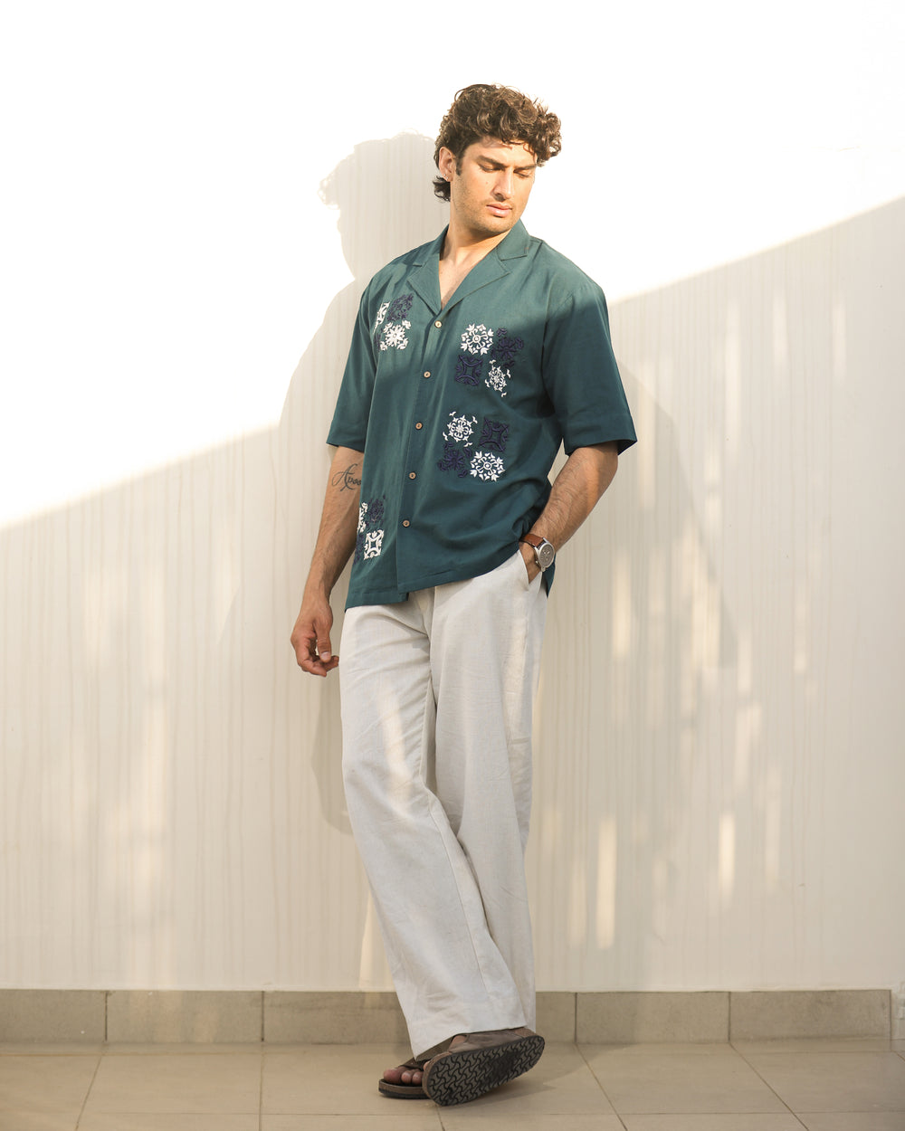 Man wearing a green shirt with floral patterns and light-colored pants against a white wall.