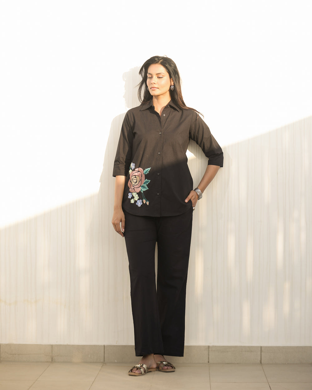 Woman wearing a black blouse with floral embroidery and black pants against a white wall.