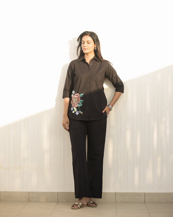 Woman wearing a black blouse with floral embroidery and black pants against a white wall.