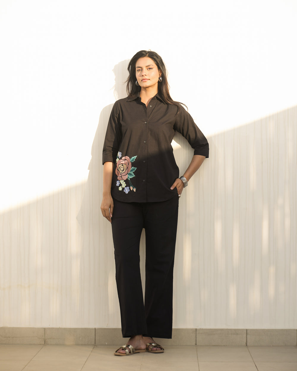 Woman wearing a black blouse with floral embroidery and black pants against a light-colored wall.